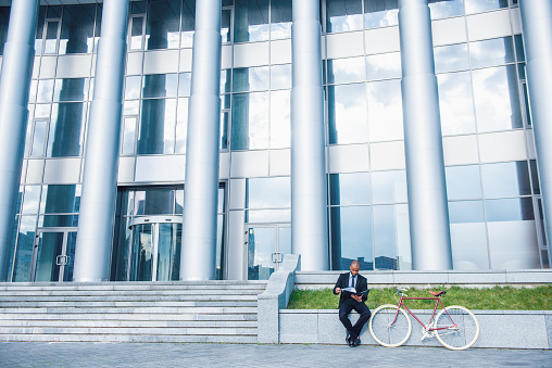 Panorama view of office building, handsome businessman in classic suit is examining documents, sitting near his bike