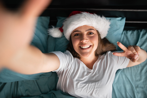 Close up photo of cheerful woman lying in bed making v-sign photos in room indoors during christmas morning. Copy space