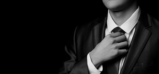 A businessman is adjusting his tie in a suit on a dark black background.