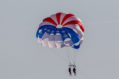 Two people parasail under a red, white, and blue parachute in front of a gray sky