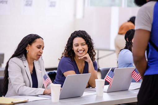 A young adult woman smiles as she trains the mature adult woman to volunteer at the polling place.