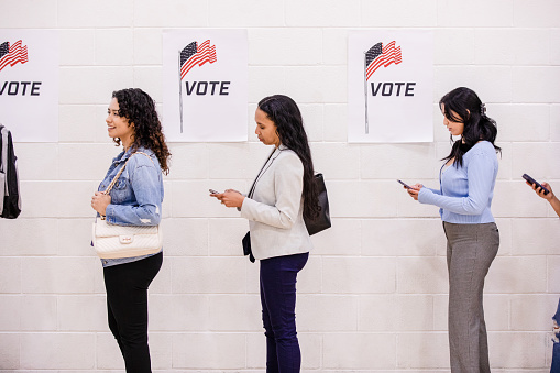 A profile view of a group of diverse women standing in line to vote.  Several of the women use their smart phones while they wait.