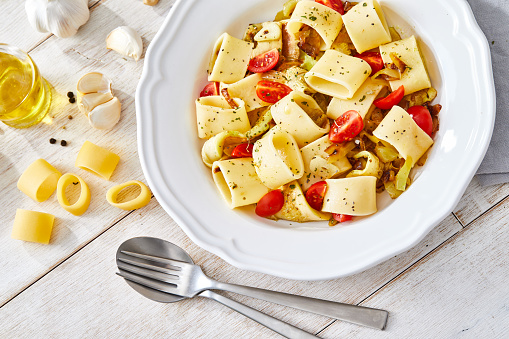 Healthy Vegan pasta, made of cherry tomato, green pepper, herbs, spices, seasoning and garlic, served on an elegant ceramic plate, with fresh vegetables and herbs, olive oil and pepper, on a white rustic wooden natural kitchen or restaurant table, representing a wellbeing and a healthy lifestyle and food indulgence, view image with a copy space