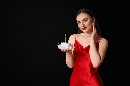 Attractive young woman holding her Birthday cake with burning candle on black background, space for text