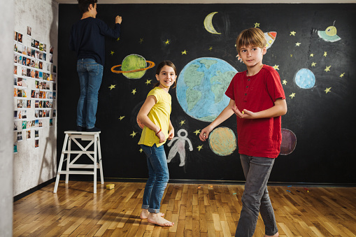 Group of children playing while drawing planets and stars with colorful chalks on the blackboard wall in the room