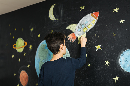 Teenage boy drawing planets and stars on a blackboard wall in the room