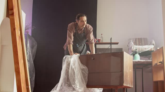 An adult Caucasian male furniture repairman in a special protective apron in a working workshop determines the scope of work on the restoration of an old chest of drawers and smiles. Advertising