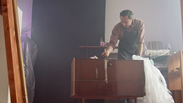 An adult Caucasian man in special clothes in a workshop removes packing polyethylene from an old chest of drawers for restoration. Small business