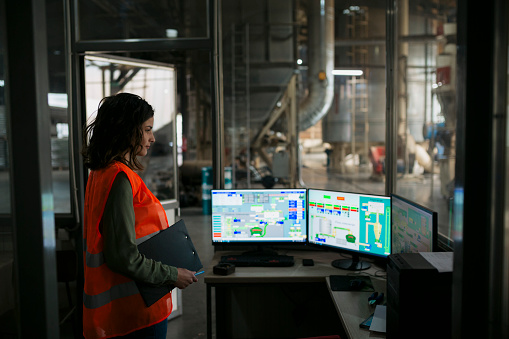 Female industrial worker operating machine on plant floor, looking at monitors over control panel. Worker looking at computer monitors showing automatic production system in wood factory.