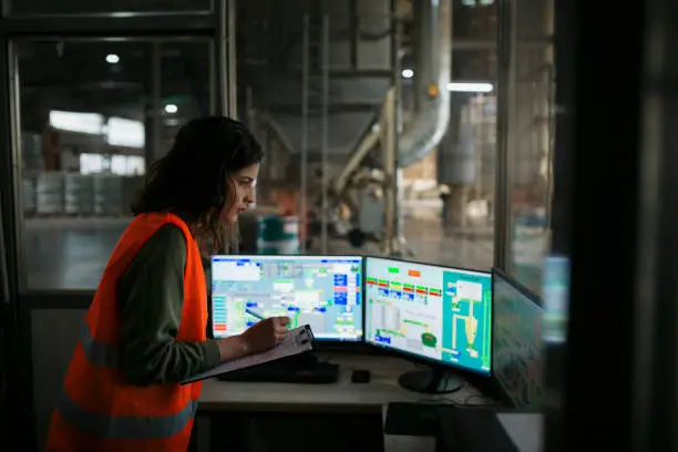 Female technician monitoring the production from the control room looking at computer monitor and making notes on clipboard. Woman wearing reflective vest making notes on clipboard while working in automated wood pellet manufacturing factory.