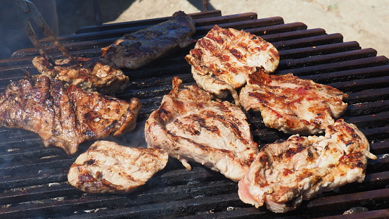 Grilled juicy meat pork steaks in burning coals on a barbecue grill, white smoke. Crispy crust. Tasty steak with blood and fried strips. Men's food. Grilled juicy striped steak is ready for a picnic.