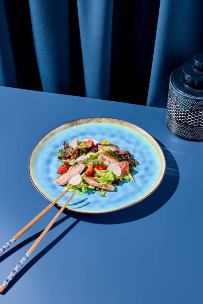 오리와 딸기로 구성된 눈에 띄는 아시아 샐러드를 파란색 세라믹 접시에 담았습니다. 드라마틱한 짙은 파란색 커튼을 배경으로 뚜렷한 그림자와 흥미로운 미니멀리즘 효과를 만들어냅니다. 현� - 16377 뉴스 사진 이미지