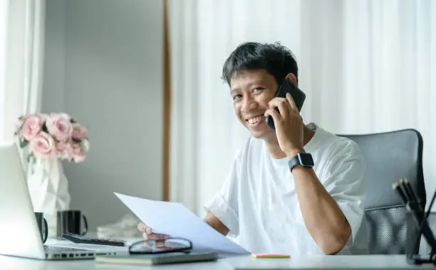 Smiling asian man sitting at workplace in modern office and talking on mobile phone.