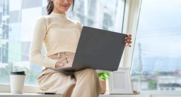 istock Smiling young woman using laptop while sitting at the large windows with cityscape. 1500754628