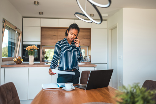 3/4 length image of a biracial woman in semi-casual clothes, picking up a paper document from the table, a coffee cup and an open laptop on the dining table where she is working. Wide angle shot.