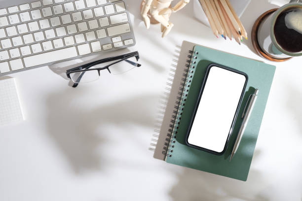 istock Smart phone with blank screen, coffee cup, notepad and keyboard on white table. Top view with copy space. 1500756183