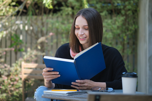 Pleased young woman in warm sweater relaxing outdoor and reading book.