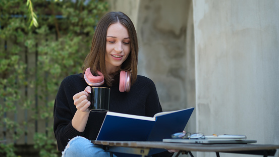 Smiling young woman in casual clothes drinking fresh hot beverage and reading book at cafe table.