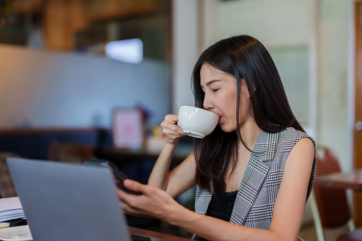 Close-up asian woman holding mobile phone with two hands, being using hand print mobile phone, chat with customers continuously, do not stop resting, sitting in bakery with cup coffee on table.