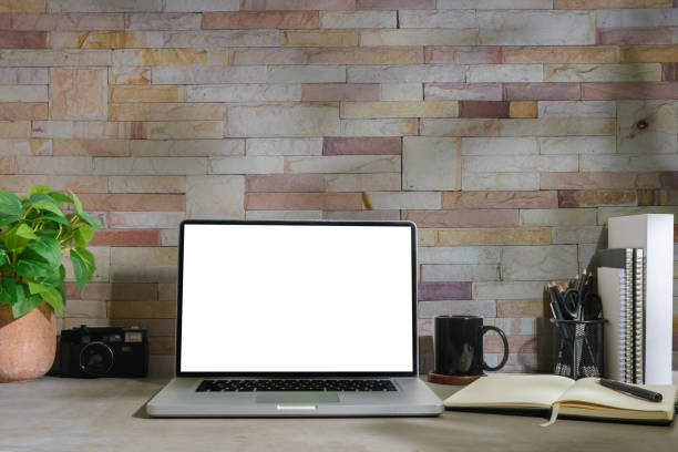 istock Front view of laptop with blank screen, coffee cup, notebook and houseplant on grey table against brick wall. 1500761110