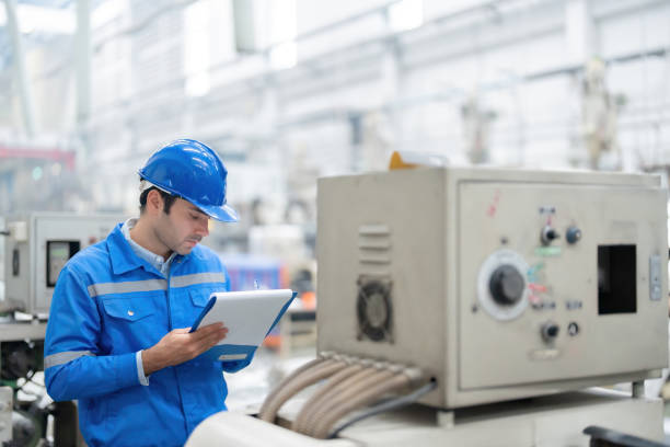 istock half body photo up american male engineer He is using a checklist pen near the control panel of a working machine, wearing a uniform and helmet, in a large plastic and metal factory. 1500764952