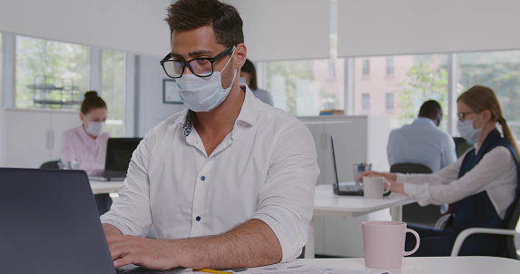 Medium shot of indian businessman in safety mask working on laptop sitting at office desk. Young male manager wearing protective mask doing financial analysis in modern office