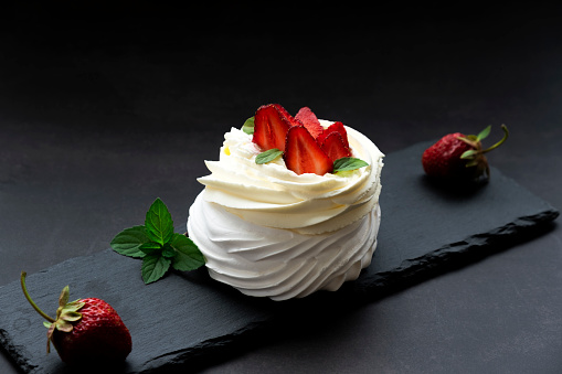 Luxury Pavlova meringue cake with fresh strawberry and whipped cream mascarpone served on black slate board on dark background. Anna Pavlova dessert for restaurant or confectionery menu.