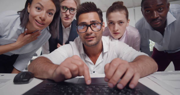 Multiethnic group of colleagues analyzing data looking at laptop screen working together in office. Indian businessman typing on computer with coworkers watching and standing behind