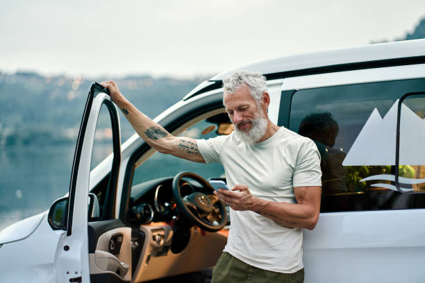 Mature man traveler standing near camper van using mobile in camping. Mature tattooed man standing near rv camper van on vacation using mobile phone. Smiling mature active traveler holding smartphone enjoying free internet in camping tourism nature park. hipster car stock pictures, royalty-free photos & images