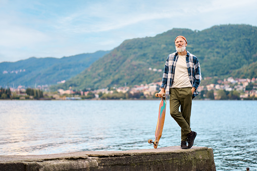 Active cool bearded old hipster man standing in nature park holding skateboard. Mature traveler skater enjoying freedom spirit and extreme sports hobby leisure lifestyle on lake background.