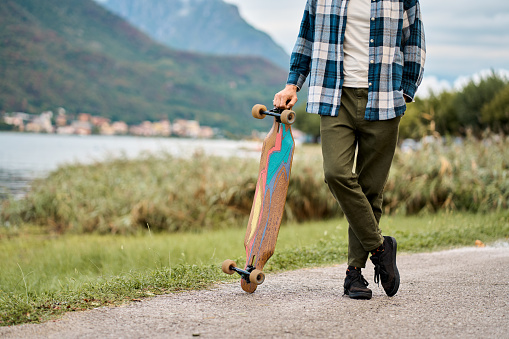 Close up view of skater healthy active man holding skateboard standing outdoors in nature park enjoying freedom spirit and extreme sports hobby with mountains view at background.