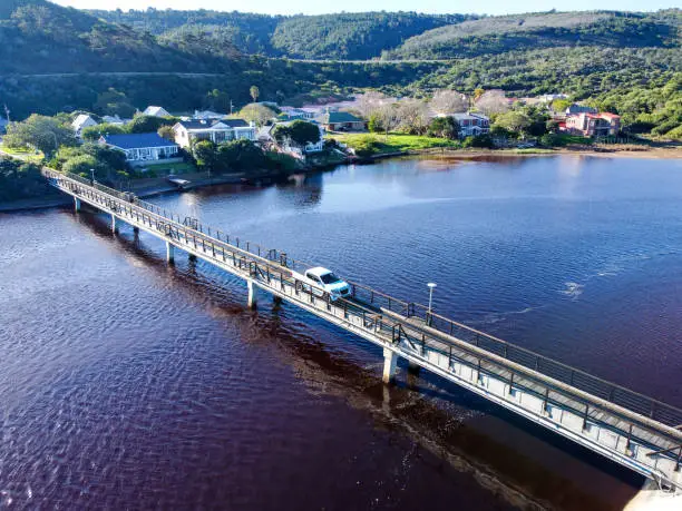 Aerial shot of a white pickup-truck driving over small wooden coastal bridge  in a dark brown lagoon and a view of green hills and green trees and bushes surrounding the bridge and various multi coloured houses on the lagoon, Die Eiland.