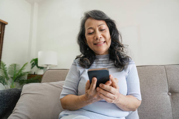 istock Smiling older asian woman sit rest on couch at home text or message online on modern cellphone gadget. Happy mature female relax browse internet on smartphone device. Elderly technology concept 1500801253