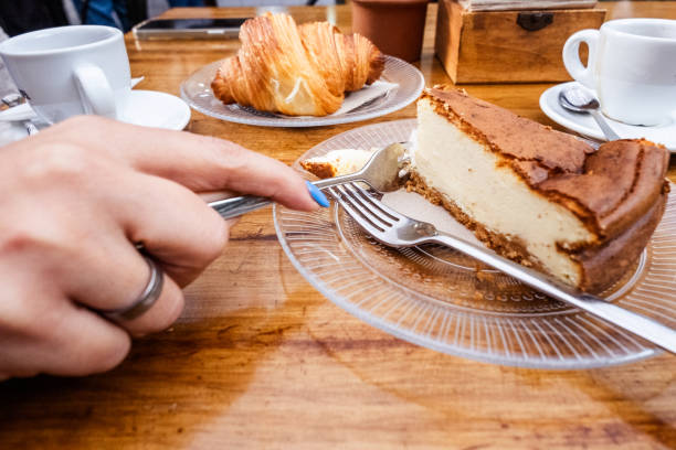Breakfast. Woman's hand with fork takes a piece of cheesecake on a glass plate, on the table there is a cup of coffee on a plate with metal spoon, glass plate with a croissant and cup of tea. All on a natural wood table. Gastronomy. Tenerife, Canary Islands, Spain