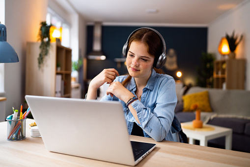 Teenage girl with headphones and laptop having online school class at home.