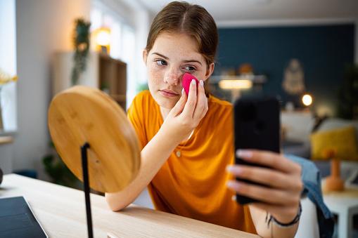 Teenage girl texting messages on her phone while doing make-up.