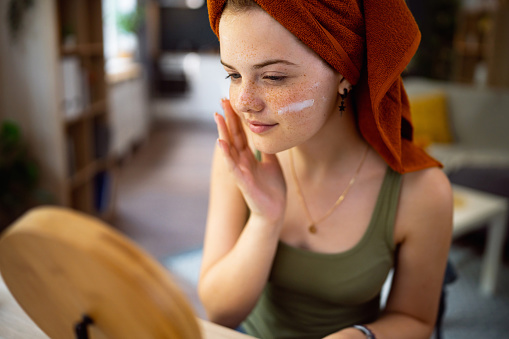 Teenage girl, with hair wrapped in a towel, applying face cream in front of the mirror.