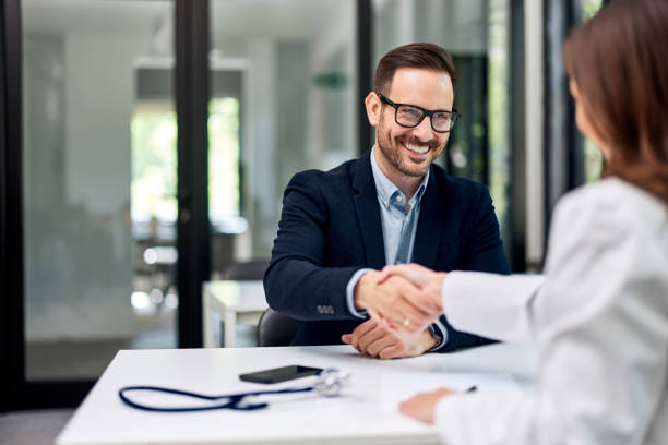 A cheerful male boss handshake with a female doctor across the office table. A smiling businessman greets a female doctor with a stethoscope on the office table and handshakes with her. doctor utensils stock pictures, royalty-free photos & images