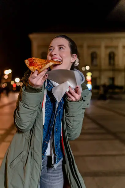 In this image, a young woman sits on a curb, enjoying a slice of pizza while looking up at the twinkling stars above