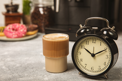 Alarm clock on the background of the coffee machine. Black alarm clock on the kitchen table against the background of a cup of aromatic coffee and a coffee machine. Cheerful morning concept. Place for