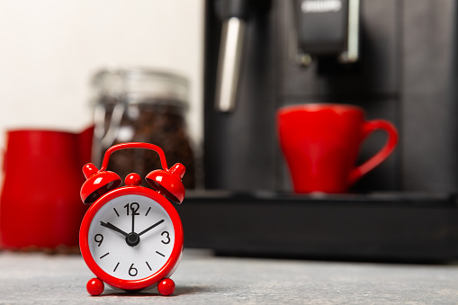 Alarm clock on the background of the coffee machine. Red alarm clock on the kitchen table against the background of a cup of aromatic coffee and a coffee machine. Cheerful morning concept.