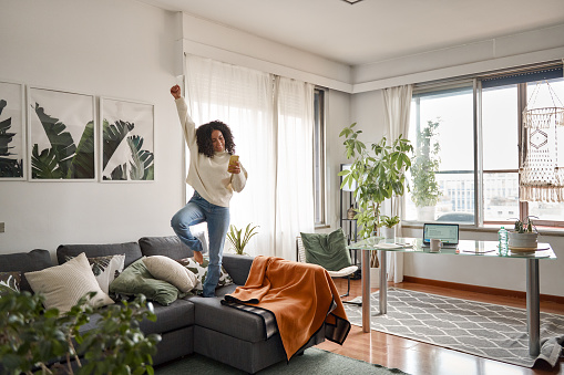 Happy excited young woman using smartphone dancing at home. Latin girl jumping on couch listening music on mobile phone, winning money in app game celebrating standing in living room interior.