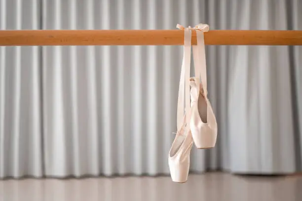 A pair of pink satin ballet pointe shoes with  satin ribbon hanging on a wooden barre in a ballet studio.