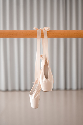 A pair of pink satin ballet pointe shoes with  satin ribbon hanging on a wooden barre in a ballet studio.