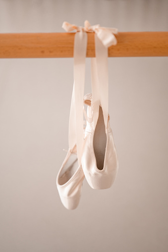 A pair of pink satin ballet pointe shoes with  satin ribbon hanging on a wooden barre in a ballet studio.