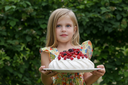 Prtrait of adorable little girl in floral summer dress holding tray with Pavlova cake