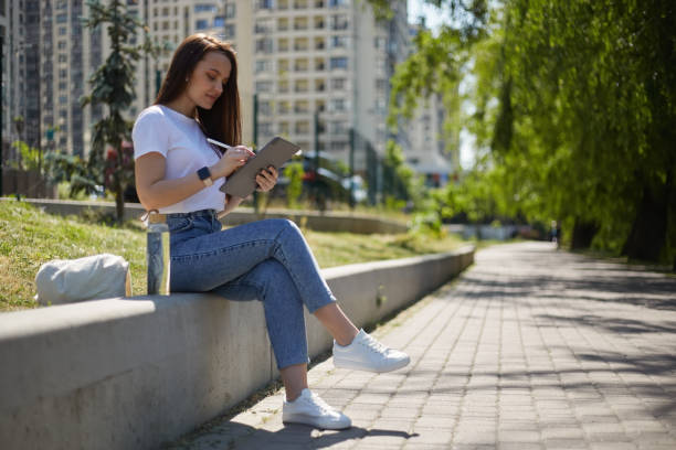 istock Young graphic designer sitting outdoor with a tablet computer and drawing a sketch with a digital pen. Illustrator female making a sketch with a stylus outdoor 1500816284