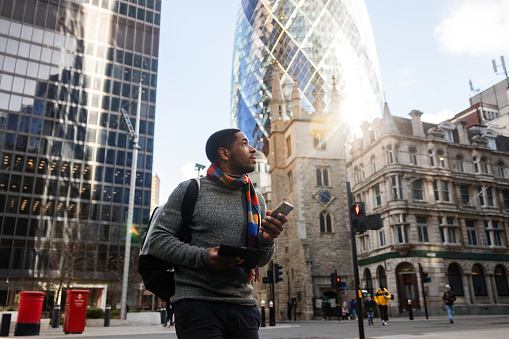 A serious adult black male on his way to a work meeting in London. He is admiring the beautiful streets of London while walking. In his hands, he is carrying a smartphone and a tablet. He looks serious. The weather is nice and sunny. He looks focused.