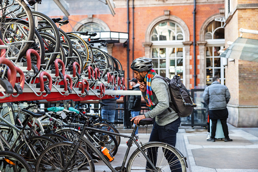 An adult black male with a helmet parking a bike in London as he arrives at his office building. He is wearing warm business casual clothes. Sustainable transport to work in London.