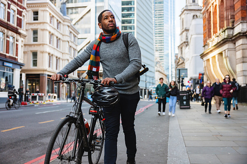 An adult black entrepreneur walking home from the office in London. He is pushing a bike while walking around the beautiful streets of London on a beautiful sunny day. The sun is illuminating the beautiful buildings in the background.